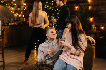Young couple talking on New Year Eve against backdrop of festive Christmas tree. Lovers boyfriend and girlfriend communicate nicely in festively decorated room. Girl holds glass of champagne in hands.