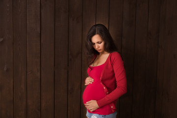 Christmas pregnant woman in jeans shorts and red top holds hands on belly on a dark brown background. Pregnancy, maternity, expectation concept. Beautiful tender mood photo of pregnancy