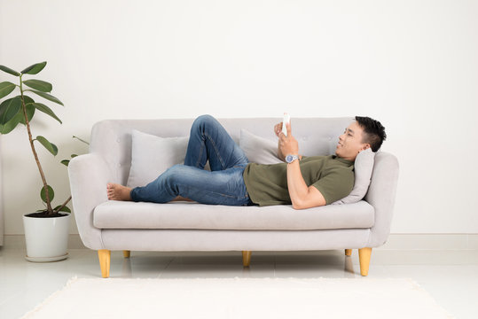 Relaxed man using a smart phone lying on an couch/sofa in the living room at home