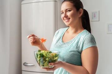 Body Care. Chubby girl standing in kitchen eating healthy salad smiling cheerful