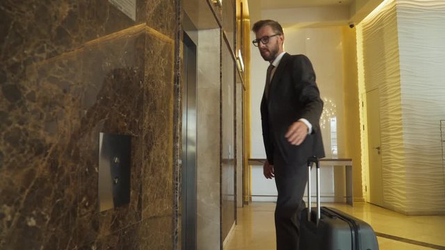 Young man in suit carrying a suitcase waiting for elevator in hotel lobby, business trip.
