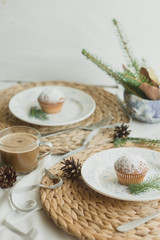 Obraz na płótnie Canvas Christmas table setting with muffins, coffee, white plates, wicker coasters, fir branches and cones. Table setting in a rustic style for the New Year.