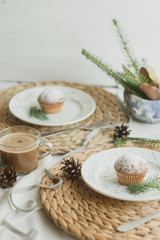 Christmas table setting with muffins, coffee, white plates, wicker coasters, fir branches and cones. Table setting in a rustic style for the New Year.