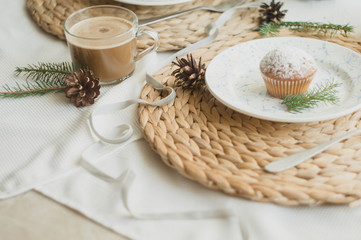 Christmas table setting with muffins, coffee, white plates, wicker coasters, fir branches and cones. Table setting in a rustic style for the New Year.