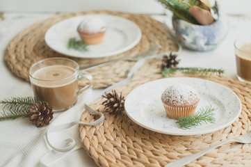 Christmas table setting with muffins, coffee, white plates, wicker coasters, fir branches and cones. Table setting in a rustic style for the New Year.