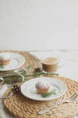 Christmas table setting with muffins, coffee, white plates, wicker coasters, fir branches and cones. Table setting in a rustic style for the New Year.