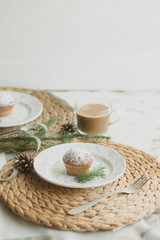 Christmas table setting with muffins, coffee, white plates, wicker coasters, fir branches and cones. Table setting in a rustic style for the New Year.