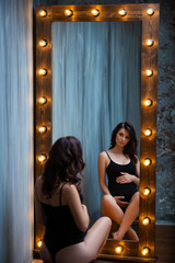 A young pregnant woman in a black bodysuit sits near a beautiful wooden mirror with lamps. Interior Loft.