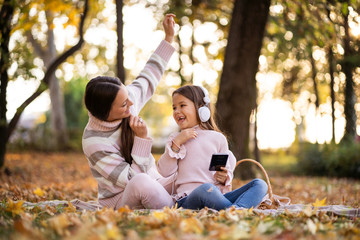 Mother and daughter enjoyng autumn in park. They are listening music on headphones.