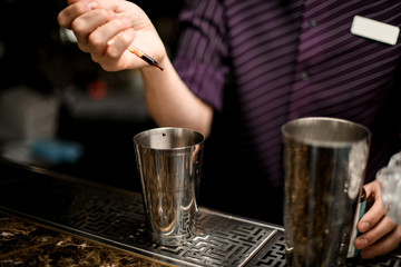Male bartender adding a brown essence with a pipette to an alcoholic drink in a steel shaker