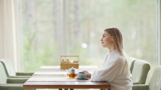 Side view zoom out shot of beautiful young woman sitting alone at cafe table, drinking tea and day dreaming with smile on her face
