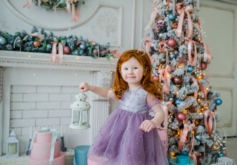 Redhead little girl in a violet dress against the backdrop of a Christmas tree with a white candlestick in the hand