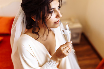 Beautiful bride in a white robe holds in her hand a glass of champagne. Elegant woman with professional make up and hair style. Wedding morning of the bride. Wedding day. Wedding dress. Fashion bride.