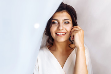 Elegant bride in white robe posing under curtain. Amazing bride in the morning preparation. Beautiful woman with professional make up and hair style. Morning of the bride. Wedding day. Fashion bride.