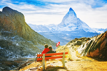 Romantic couple viewing iconic mountain peak Matterhorn, tourist young couple resting on wooden bench with majestic view at famous tourist landmark - Matterhorn enjoying of recreation mountain trip.