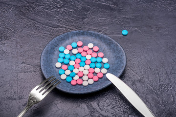 Obraz na płótnie Canvas a bunch of colorful pills on a plate of dark color, next to a fork and a knife and it's all on a dark background