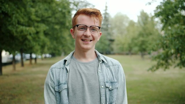 Slow motion of handsome redhead guy wearing glasses smiling in camera in green ity park in summer. Happy people, nature and cheerful youth concept.