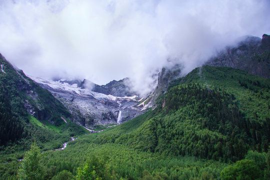 Beautiful panorama of the Caucasus mountains. Top of mountain range is covered in fog. Clouds lie on mountains. Mountain river. Summer day. Background image for travel and nature.