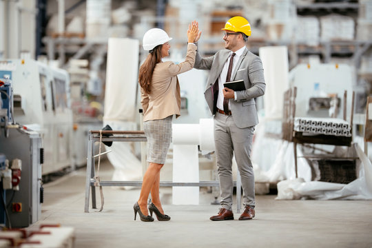 Businessman and businesswoman in factory. Man and woman in suits with helmets in factory giving each other high five. - Powered by Adobe