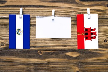 Hanging flags of El Salvador and Gibraltar attached to rope with clothes pins with copy space on white note paper on wooden background.Diplomatic relations between countries.