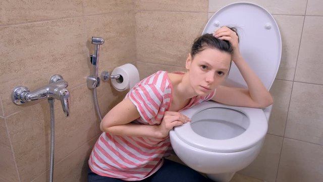 Drunk tired woman is vomiting in toilet sitting on the floor at home and looking at camera, morning hangover, side view.