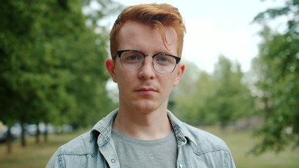 Slow motion of serious young man in glasses standing alone in green park in city and looking at camera. Emotions, youth and summer nature concept.