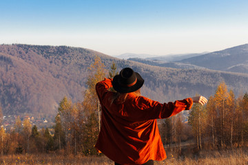 Fototapeta na wymiar Happy and free girl or woman in a brown velvet shirt and hat is dancing against the backdrop of the mountains. Amazing mountain view with yellow trees and blue hills.
