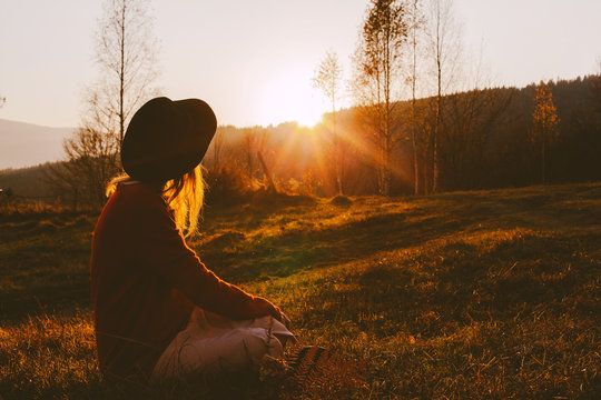 A Dreamy Young Girl Or Woman Traveler In A Fedora Hat Sits In The Grass And Enjoys The Mountain View, The Warm Rays Of The Autumn Sun And A Beautiful Sunset.