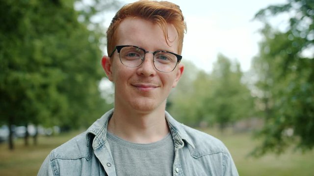 Portrait of handsome male student starting to smile standing in city park alone looking at camera. Mood change, human emotions and young people concept.