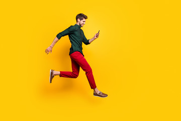 Fototapeta na wymiar Full length profile side photo of positive cheerful guy use smartphone search discounts on social media jump run fast hurry wear green shirt red pants trousers isolated yellow color background