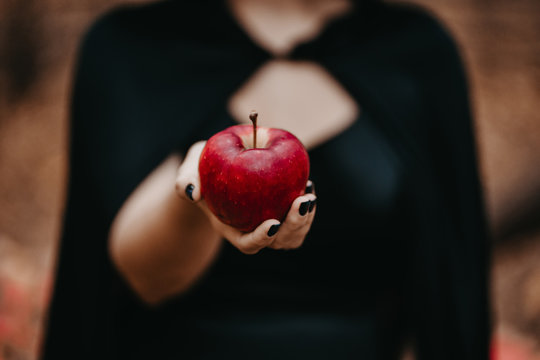 Woman as witch in black offers red apple as symbol of temptation, poison. Fairy tale concept, halloween, cosplay.