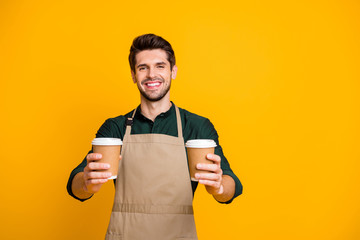 Portrait of positive cheerful guy worker in coffee bar shop hold paper card cup offer to his client mug with hot cappuccino wear apron green shirt isolated over yellow color background