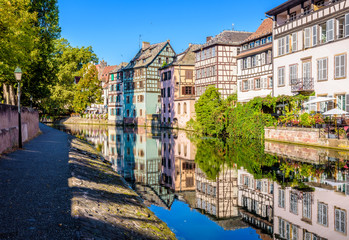 The half-timbered houses lining the canal of the river Ill in the Petite France quarter in Strasbourg, France, reflect in the still waters under a bright sunshine. - obrazy, fototapety, plakaty