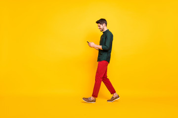 Fototapeta na wymiar Full length body size photo of moving cheerful walking addicted attractive man browsing through his telephone wearing red pants trousers footwear isolated over yellow vivid color background