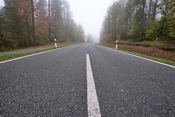 Diminishing perspective of a lonesome empty countryside road with delineators and a bicycle path along leading through a forest with dense fog. Seen in October in Germany in Franconia / Bavaria.