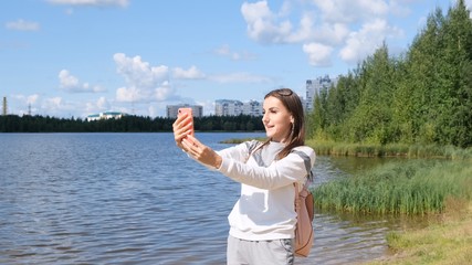 Beautiful girl with a backpack takes a selfie near the lake and talks on a cell phone on Skype.