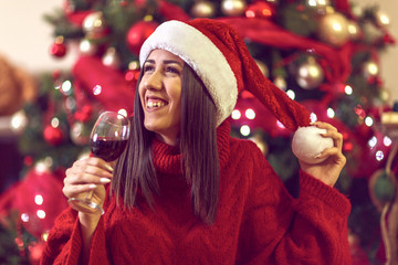 young woman in Santa hat by Christmas tree at home toasting with red wine.