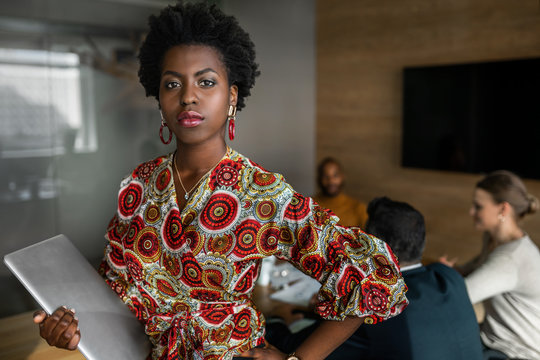 Beautiful young confident professional black african business woman holding laptop, coworkers hold a meeting in background
