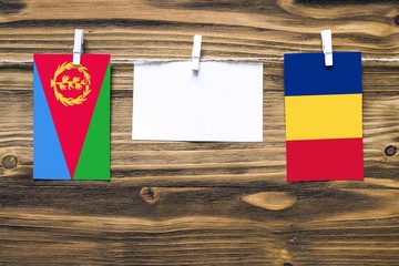 Hanging flags of Eritrea and Romania attached to rope with clothes pins with copy space on white note paper on wooden background.Diplomatic relations between countries.