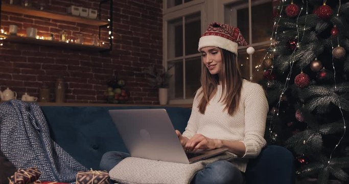 Beautiful Young Woman Wearing Santas Hat Sitting near Christmas Tree at Home Background Using her Laptop Chatting Texting Concept of Holidays and New Year Side View.