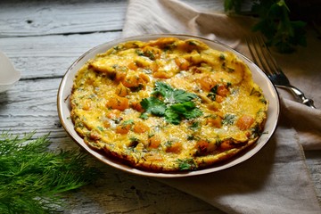 Omelet in a white beautiful plate. Omelet with yellow tomato, pepper and parsley leaves. Appetizing breakfast. Wooden light background. Egg shells, textiles.