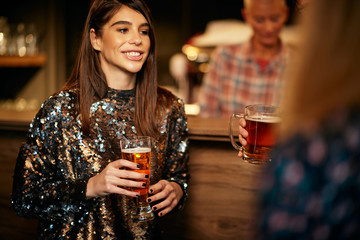 Beautiful caucasian smiling stylish woman  holding beer and chatting with friends. In background is bar counter. Pub interior.