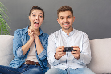 Relationnship Concept. Young couple man playing video game and woman looking at tv sitting on sofa studio isolated on grey smiling cheerful