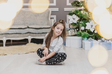 Angelic child daughter is sitting in lotos position on the carpet near glittering Christmas tree....