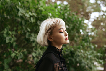 Young adult blond asian female in dark clothes with her eyes closed outdoors, selective focus