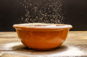 brown bowl stands on a wooden table against a black background, flour is poured into the bowl. Close-up photo