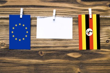 Hanging flags of European Union and Uganda attached to rope with clothes pins with copy space on white note paper on wooden background.Diplomatic relations between countries.