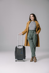 beautiful smiling girl in beige coat posing with travel bag on grey