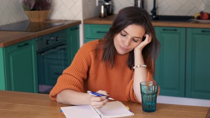 Beautiful young woman making notes sitting in kitchen, 4K