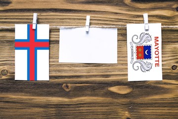 Hanging flags of Faroe Islands and Mayotte attached to rope with clothes pins with copy space on white note paper on wooden background.Diplomatic relations between countries.